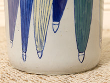 Load image into Gallery viewer, Close-up of a ceramic container or vase with a white glaze and a repeating hand-painted pattern of stylized umbrella-like shapes in blue, pale yellow, and green, resting on a woven beige mat.