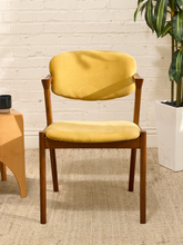Load image into Gallery viewer, Image 73 of 89: A mid-century modern wooden dining chair with a mustard yellow velvet upholstered seat and backrest, set against a white brick wall background.