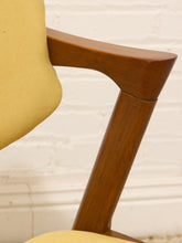 Load image into Gallery viewer, Image 80 of 89: Close-up of a mid-century modern chair featuring a warm brown wooden frame and a mustard yellow fabric seat and back cushion, set against a white brick wall background.