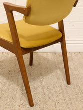 Load image into Gallery viewer, Image 87 of 89: A close-up of a mid-century modern wooden armchair with mustard yellow velvet upholstery, positioned on a textured beige woven rug against a white brick wall.