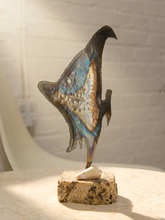 Load image into Gallery viewer, A sculptural fish ornament with an iridescent blue, gold, and bronze finish, mounted on a textured stone base with a small white shell accent, set against a softly blurred white brick wall background.
