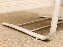 Load image into Gallery viewer, Close-up of a white painted metal chair leg resting on a beige woven rug against a white wall baseboard.