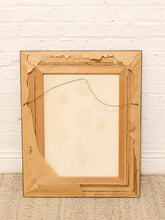 Load image into Gallery viewer, The back of a vintage wooden picture frame, showing the mounting board, wire hanger, and worn edges, leaning against a white brick wall on a woven rug.
