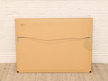 Load image into Gallery viewer, The back of a large square canvas or framed art print, showing a tan cardboard backing, metal hanging hardware, and a wire support system, leaning against a white brick wall on a woven rug.