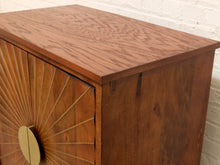 Load image into Gallery viewer, A close-up of a mid-century modern wooden cabinet with a warm brown wood finish, featuring a sunburst inlay design on the front door and a round brass pull handle, set against a white brick wall background.