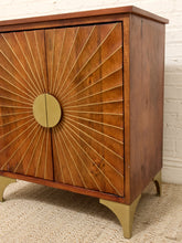 Load image into Gallery viewer, Mid-century modern wooden cabinet with sunburst metal detailing on the doors and brass legs, set against a white brick wall and beige rug.