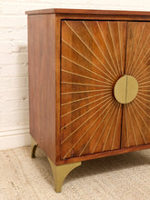 Load image into Gallery viewer, Mid-century modern wooden cabinet with brass sunburst detailing on the doors and matching brass legs, shown in a styled room setting against a white brick wall and beige rug.