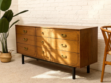 Load image into Gallery viewer, A mid-century modern wooden dresser with six drawers, brass handles, and black legs, set against a white brick wall with a potted plant on the left and a wooden chair on the right.