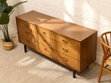 Load image into Gallery viewer, A mid-century modern wooden sideboard with six drawers, brass handles, and black legs, styled in a room with a white brick wall, beige rug, and a potted plant.