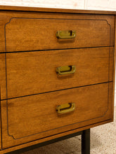 Load image into Gallery viewer, A close-up of a mid-century modern wooden chest of drawers with three drawers, featuring a warm medium-brown wood grain finish, black painted trim, and brass handles.
