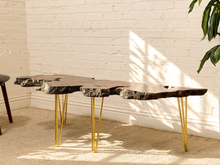 Load image into Gallery viewer, A modern live-edge wood console table with a dark natural bark top and slim gold metal hairpin legs, set against a white brick wall with warm sunlight and leaf shadows.
