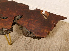 Load image into Gallery viewer, A rustic live-edge wooden coffee table with a rich, dark brown burl wood top and gold metal legs, shown on a textured beige rug against a white brick wall.