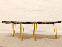 Load image into Gallery viewer, A modern coffee table with a dark, textured live-edge stone top and gold metal hairpin legs, set against a white brick wall and beige carpet.