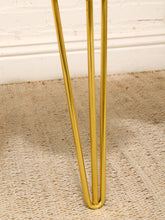 Load image into Gallery viewer, Close-up of polished gold metal hairpin table legs standing on a beige woven rug against a white brick wall.