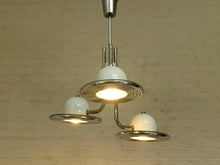Load image into Gallery viewer, A three-light pendant ceiling fixture with a polished silver metal frame and white glass shades, shown hanging against a light beige brick wall.