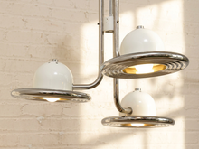 Load image into Gallery viewer, A mid-century modern three-light chandelier with glossy white dome-shaped glass shades and polished chrome metal arms and rings, shown illuminated against a white brick wall.