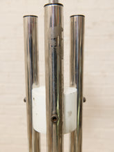 Load image into Gallery viewer, A close-up of a vintage mid-century modern metal chandelier or lamp fixture with four polished chrome tubes and a white cylindrical diffuser or base, set against a light beige brick wall.