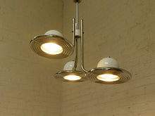 Load image into Gallery viewer, A three-light chandelier with a polished chrome metal frame and white dome-shaped glass shades, shown hanging against a white brick wall.