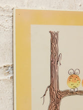 Load image into Gallery viewer, A close-up of a framed or mounted art print showing a whimsical illustration of a small orange-yellow owl perched on a brown tree branch, with a pale cream center panel and a wide warm yellow border, photographed against a textured off-white wall.