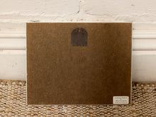 Load image into Gallery viewer, The back of a square brown textured art print or card, featuring a small dark arch-shaped cutout near the top and a small white label in the bottom right corner, leaning against a white brick wall on a woven rug.