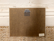 Load image into Gallery viewer, The back of a square brown art print or framed artwork, showing a metal hanger at the top and a small white label in the lower right corner, leaning against a white paneled wall on a woven rug.