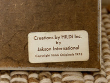 Load image into Gallery viewer, Close-up of a textured brown paperboard or wood block with a cream-colored label that reads 'Creations by HILDI Inc. by Jakson International Copyright Hildi Originals 1973', resting on a woven beige rug.