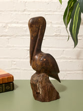 Load image into Gallery viewer, A hand-carved dark brown wooden pelican sculpture standing on a rustic wood base, displayed on a green tabletop against a white brick wall with a plant leaf in the upper right.
