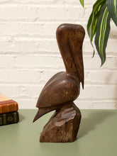 Load image into Gallery viewer, A dark brown carved wooden pelican sculpture standing on a rough tree stump base, displayed on a muted green tabletop against a white brick wall with a green plant leaf in the upper right and books on the left.