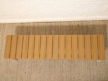 Load image into Gallery viewer, A rectangular wooden bench with vertical slats in a light natural wood finish, placed on a textured beige rug against a white brick wall.
