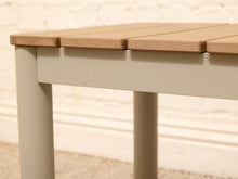 Load image into Gallery viewer, Close-up of a slatted wooden table or bench with a light beige wood top and a muted gray-beige frame, shown against a soft white brick wall background.
