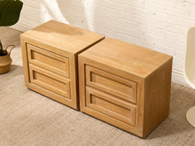 Load image into Gallery viewer, Two light oak wood nightstands with three drawers each, shown side-by-side on a beige carpet against a white brick wall.