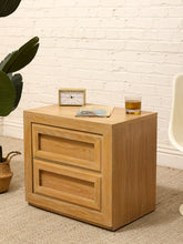 Load image into Gallery viewer, A light oak wood two-drawer nightstand shown in a styled room setting with a white brick wall, a potted plant, and a chair, topped with a small clock, a magazine, and a glass of whiskey.