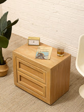 Load image into Gallery viewer, A light oak wood two-drawer nightstand styled in a room setting with a white brick wall, beige carpet, a potted plant, and a white chair. The nightstand is styled with a small clock, a magazine, and a glass of whiskey.