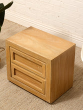 Load image into Gallery viewer, A light oak wood two-drawer nightstand with a warm honey finish, shown in a styled room setting on a textured beige carpet against a white brick wall.