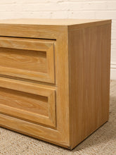 Load image into Gallery viewer, A close-up of a light oak wood nightstand or end table with two drawers, featuring a warm honey-toned finish and a visible wood grain pattern, set against a white brick wall and beige carpet.