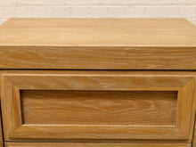 Load image into Gallery viewer, Close-up of a light oak wood nightstand or dresser drawer front with a recessed panel design, set against a white painted brick wall.