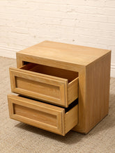 Load image into Gallery viewer, A light oak wood nightstand with two pull-out drawers, shown on a textured beige carpet against a white painted brick wall.