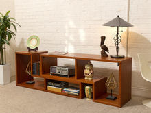 Load image into Gallery viewer, A wooden media console with open shelves and side cubbies, styled with books, a radio, vases, a bird figurine, and a table lamp in a bright room with a white brick wall.