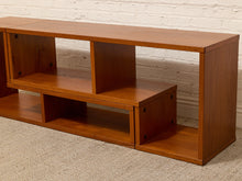 Load image into Gallery viewer, A wooden TV stand or console shelf with multiple open compartments, shown in a warm medium-brown finish against a white brick wall and light carpet.