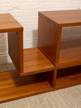 Load image into Gallery viewer, A close-up of a mid-century modern wooden TV stand or console table with a warm reddish-brown finish, featuring geometric open cubby shelves, set against a white painted brick wall and light beige carpet.