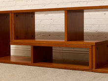 Load image into Gallery viewer, A close-up of a wooden shelving unit or media console with open cubbies, shown against a white brick wall and carpeted floor.