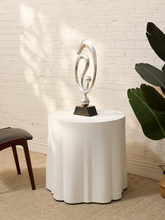 Load image into Gallery viewer, A modern silver abstract sculpture on a black base, displayed on a white pedestal table in a room with a white brick wall, beige carpet, and a green plant.
