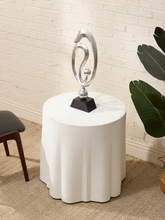 Load image into Gallery viewer, A modern silver abstract sculpture on a black base, displayed on a white cylindrical side table in a styled room setting with a white brick wall, beige rug, and green plant.