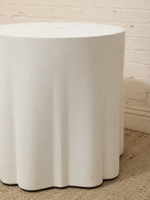 Load image into Gallery viewer, A white cylindrical side table with a fluted, scalloped edge and a textured fabric-like surface, set on a beige carpet against a white brick wall.