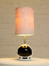 Load image into Gallery viewer, A table lamp with a cylindrical textured fabric shade, a gold-finished stem, and a glossy white and black astronaut helmet-shaped base, shown lit against a white brick wall background.