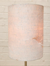 Load image into Gallery viewer, A cylindrical table lamp shade with a textured, distressed off-white linen finish, glowing with a warm light from within, set against a white painted brick wall.