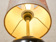 Load image into Gallery viewer, Close-up of a lamp with a warm glowing light bulb inside a beige fabric shade, showing a brass-toned metal socket and support rods.