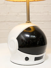 Load image into Gallery viewer, A table lamp shaped like a white astronaut helmet with a glossy black visor, a gold metal stem, and a warm yellow lampshade, set against a white brick wall background.