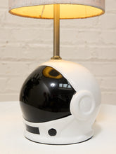 Load image into Gallery viewer, A table lamp shaped like a white astronaut helmet with a glossy black visor, a gold-toned stem, and a light beige drum shade, set against a white brick wall background.
