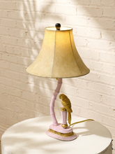 Load image into Gallery viewer, A pink and gold table lamp featuring a curved pink stem base with a gold bird figurine, topped with a beige fabric lampshade, set on a white round table against a white brick wall.